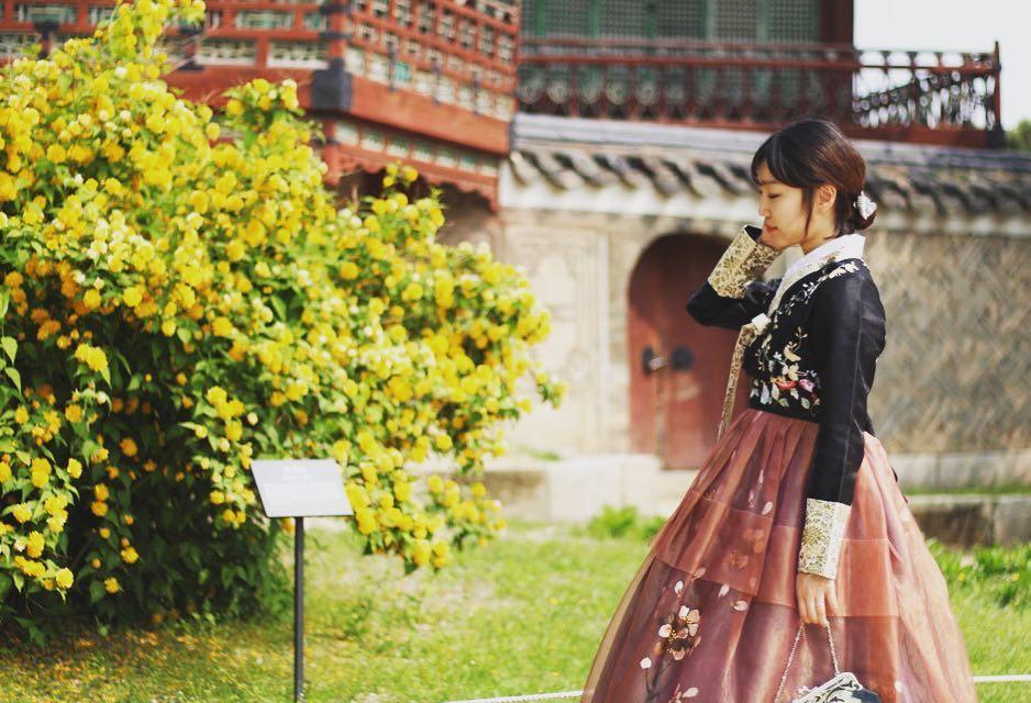 A woman wearing traditional Korean hanbok at 경복궁 (Gyeongbokgung Palace) with plants and temples in the background.