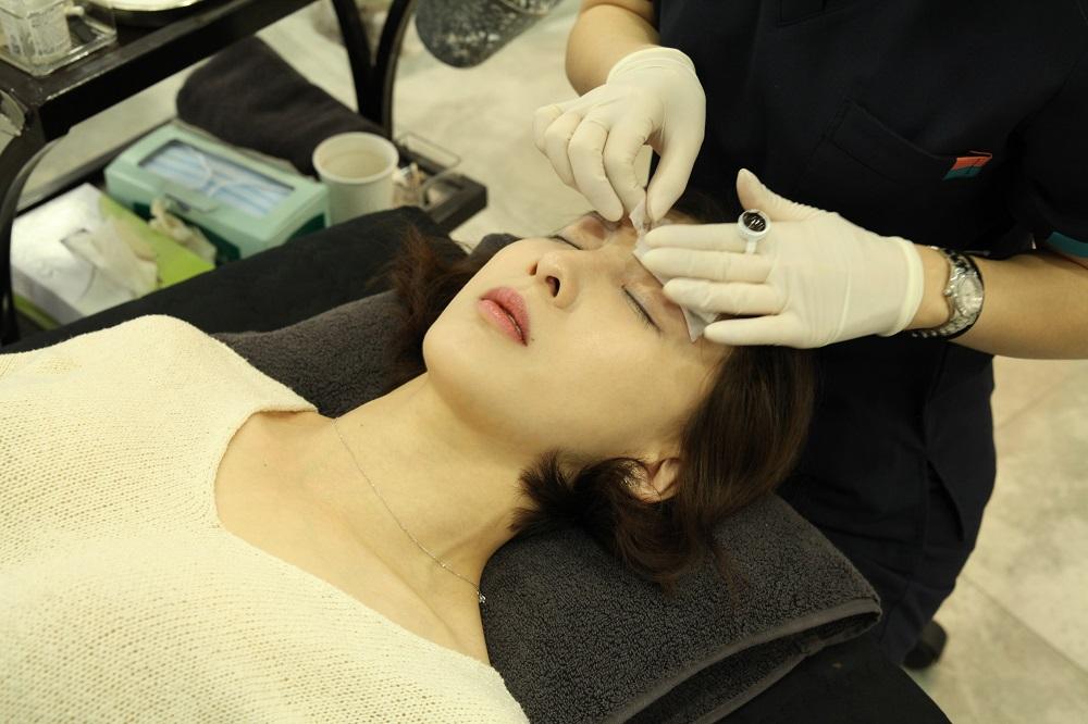 Beauty salon client receiving micropigmentation on eyebrows while hand and lip gesture showcase comfort.