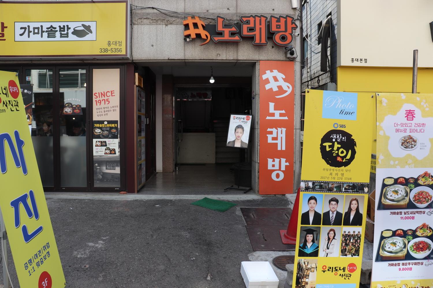 Exterior of 우리동네사진관 홍대점 in Korea showcasing fixtures, advertising posters, signage, logo, and a vending machine near the entrance.