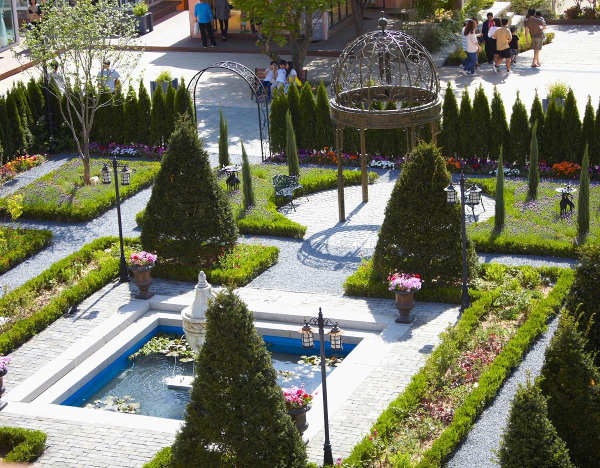 Manicured formal garden with fountain pool, trimmed cone shrubs, gravel paths, metal gazebo and visitors nearby
