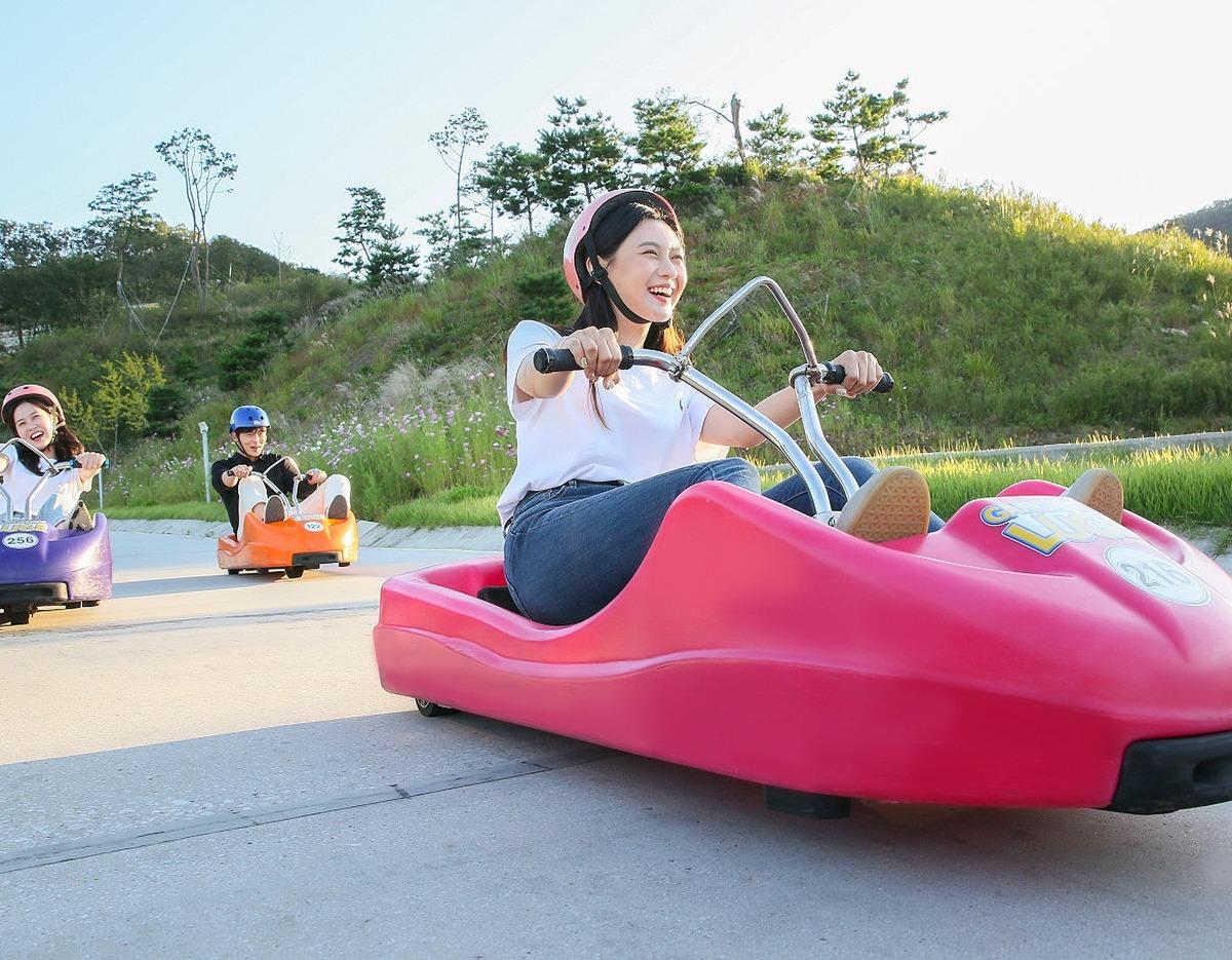 Woman laughing while riding a bright pink luge cart on a paved track with others and grassy hills behind