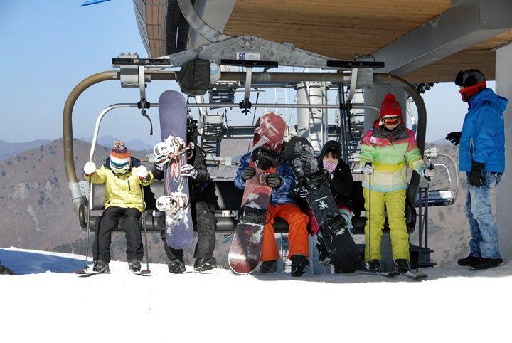 Children and instructor seated on a ski lift at Eden Valley holding snowboards and skis, snowy slope and mountains behind