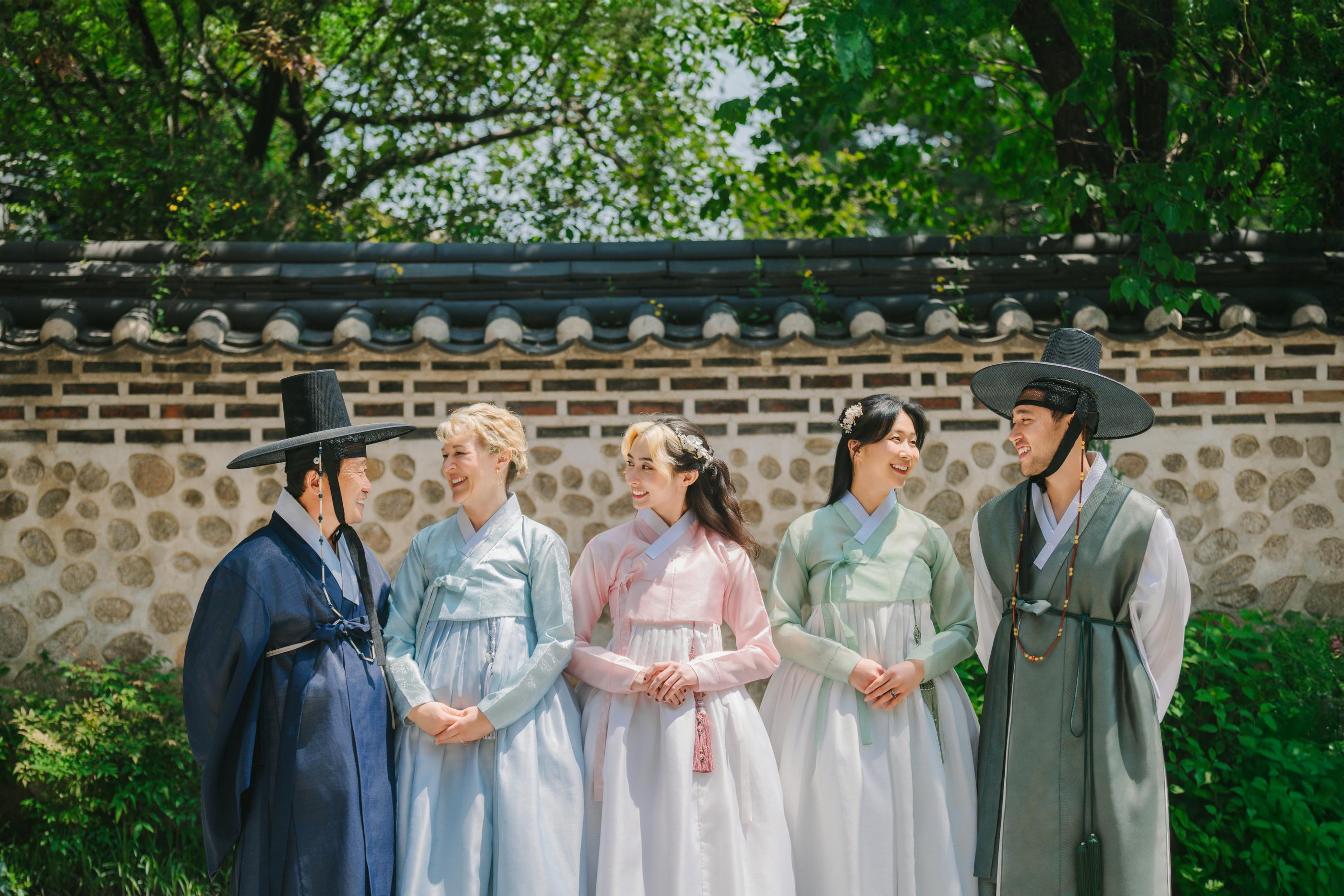 Five people in colorful hanbok standing in front of a traditional stone wall and tiled roof at Bukchon Photo Studio.