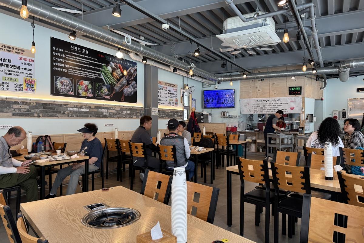 Casual Korean seafood restaurant interior with wooden tables, grilling stations, diners eating, and menu boards on walls.