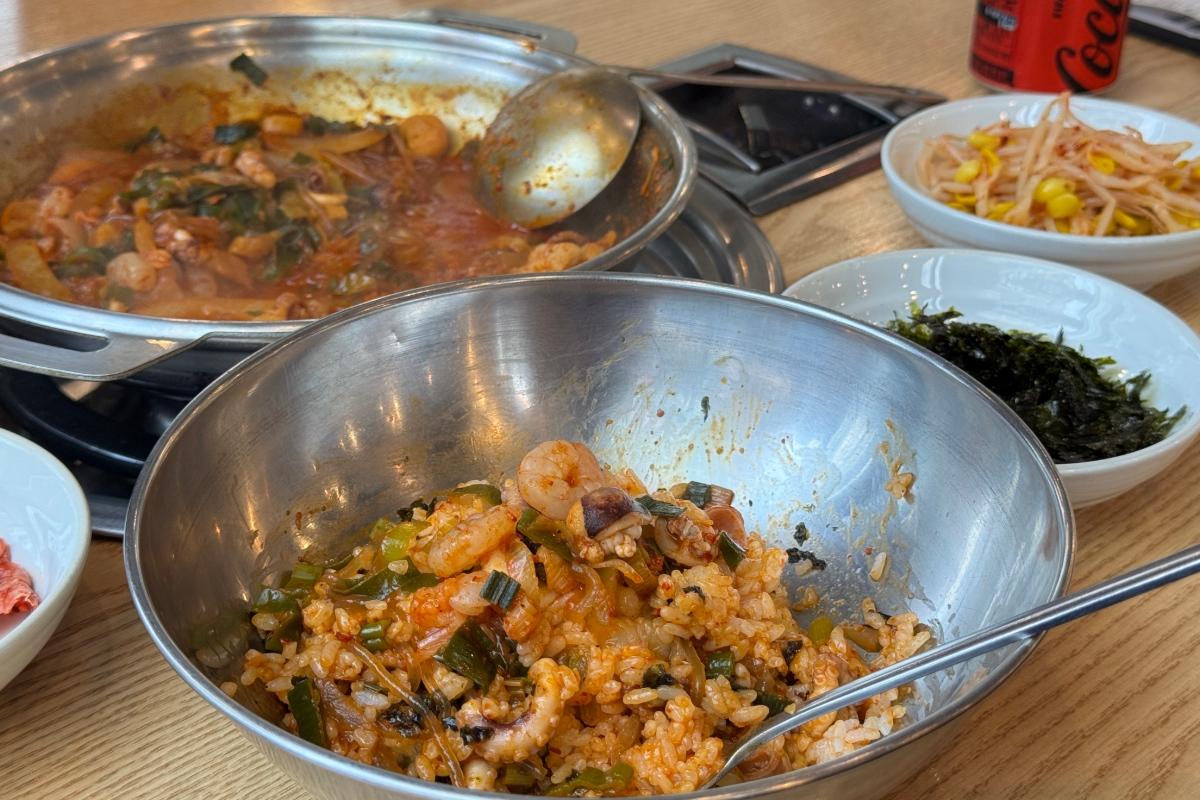 Century Octopus mixed rice with small octopus, shrimp, scallions and spicy sauce in metal bowl with side dishes and stove pot behind