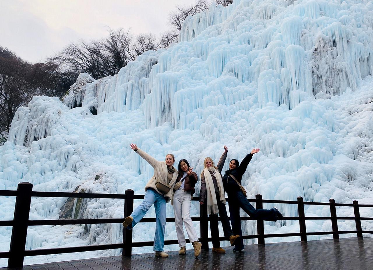 Four women posing and cheering on a wooden viewing deck in front of the towering blue-white ice wall at Eobi Ice Valley.