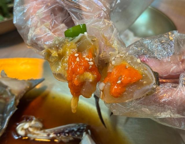 Hands in plastic gloves holding split soy-marinated crab showing bright orange roe, sesame seeds, and sauce dripping into metal dish