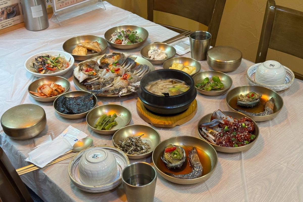 Table filled with Korean banchan and central soy-marinated crab platter, soup pot, rice bowls and metal cups at Soy Sauce Crab Restaurant