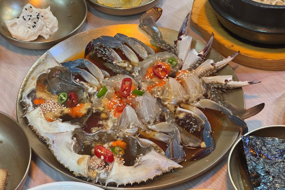 Soy-marinated raw blue crab halved and arranged on a brass plate, topped with sesame seeds, chili slices and roe, with side dishes visible