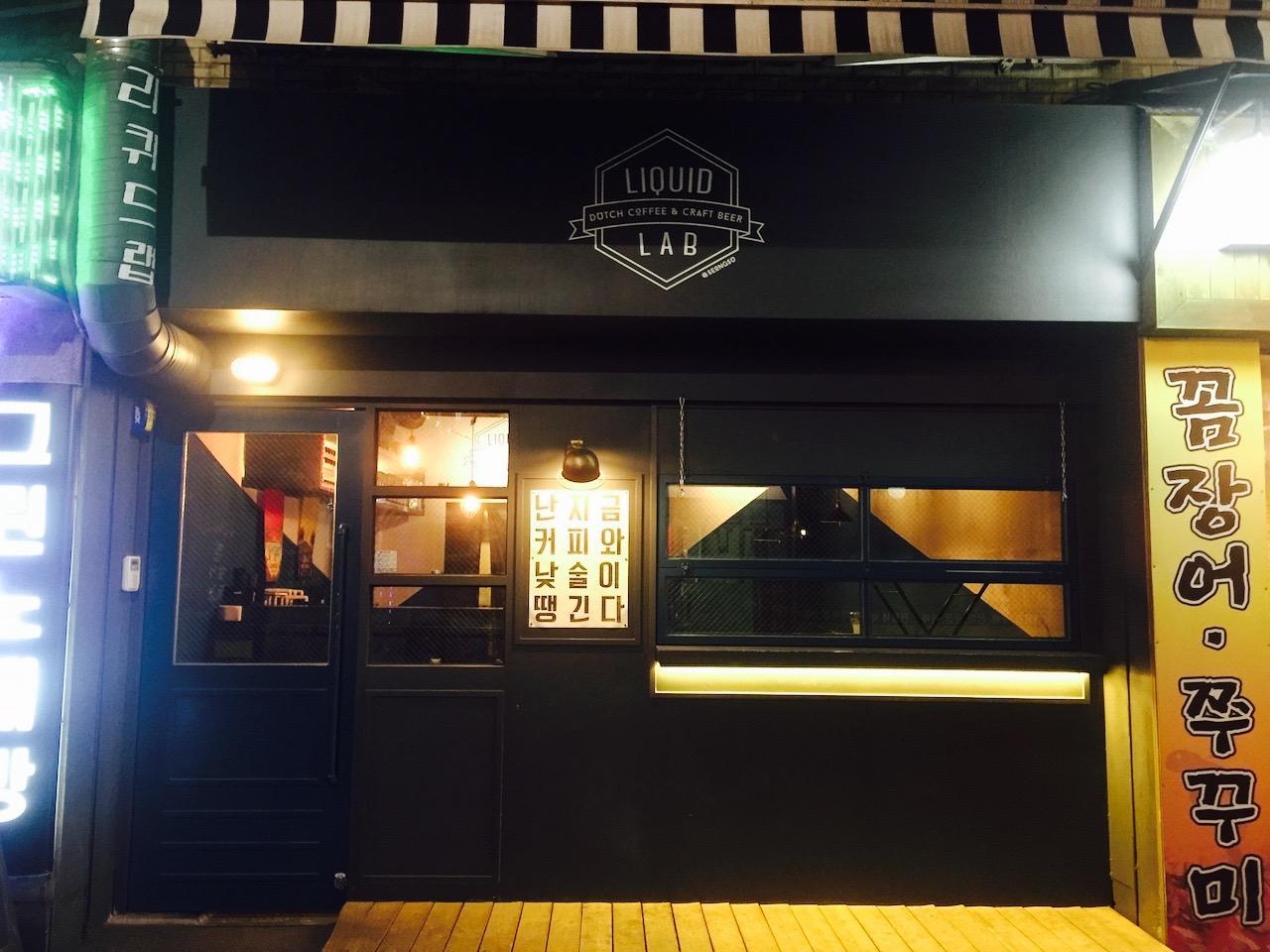 A yellow liquid lab building in Seoul, with signage for a beer pub, featuring a logo and door with machine font. The facade is illuminated at night.
