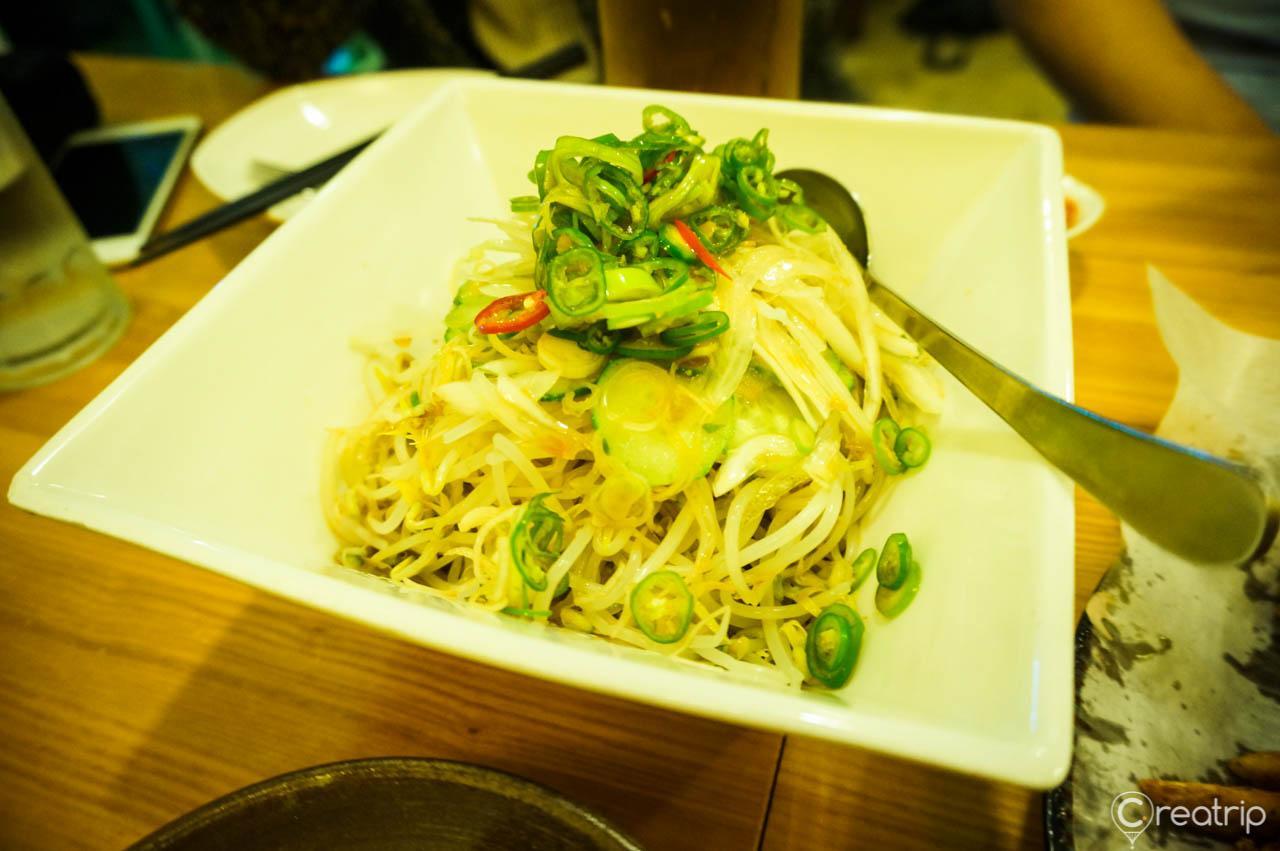 Delicious Korean cuisine featuring Saichi chicken, salad, noodles with al dente texture, and leafy greens. Served with tableware and ingredients for a perfect meal.