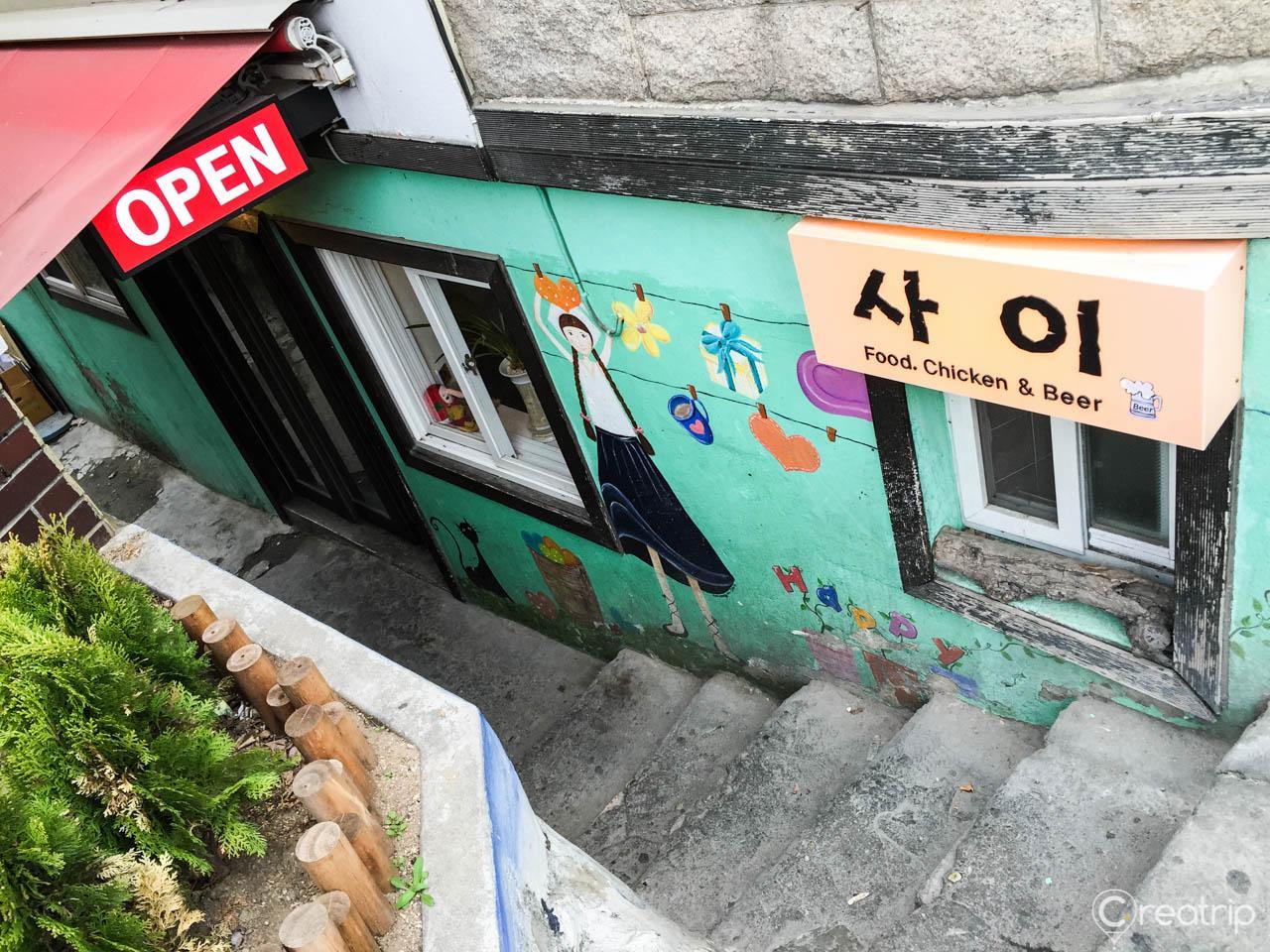 Colorful graffiti art on a wall in front of Saichi Chicken, a popular eatery in Buam-dong, Korea, with a plant and asphalt background.