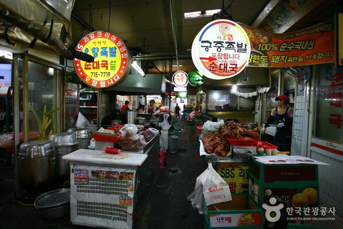 A bustling retail market in Seoul's 공덕족발골목 (Gongdeok Jokbal Alley), with hawker stalls selling goods and customers perusing the diverse offerings.