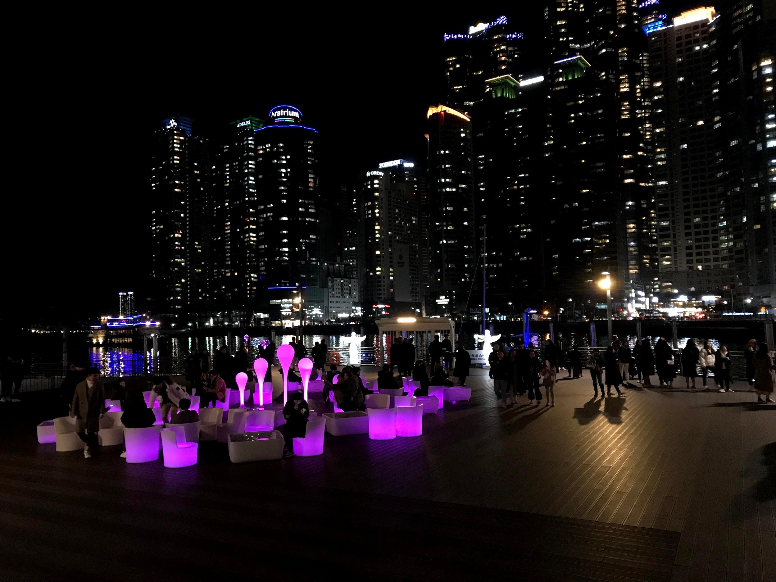 Cityscape of Busan featuring the iconic skyscraper, The Bay 101, surrounded by purple skies and water. Tower blocks, plants and electricity are visible.