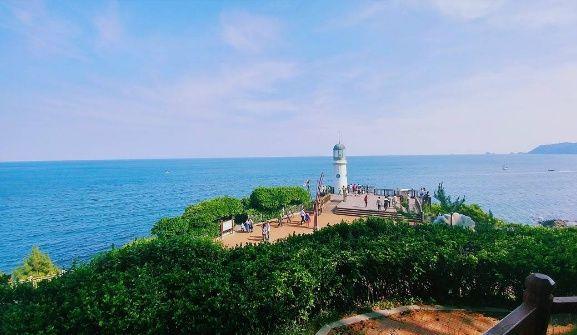 Scenic view of Dongbaek Island's coastal path, lighthouse, and tower against blue skies and water.