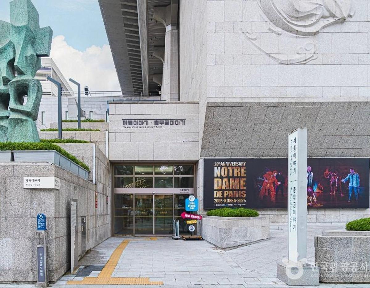 Entrance to SBS Deungchon Public Hall with stone facade, promotional poster for Notre Dame de Paris and abstract sculpture nearby