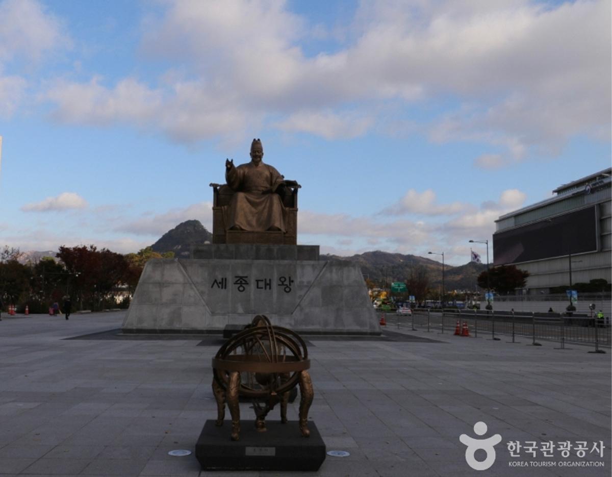 Bronze statue of King Sejong seated on a stone pedestal at Gwanghwamun Square with mountains and plaza behind