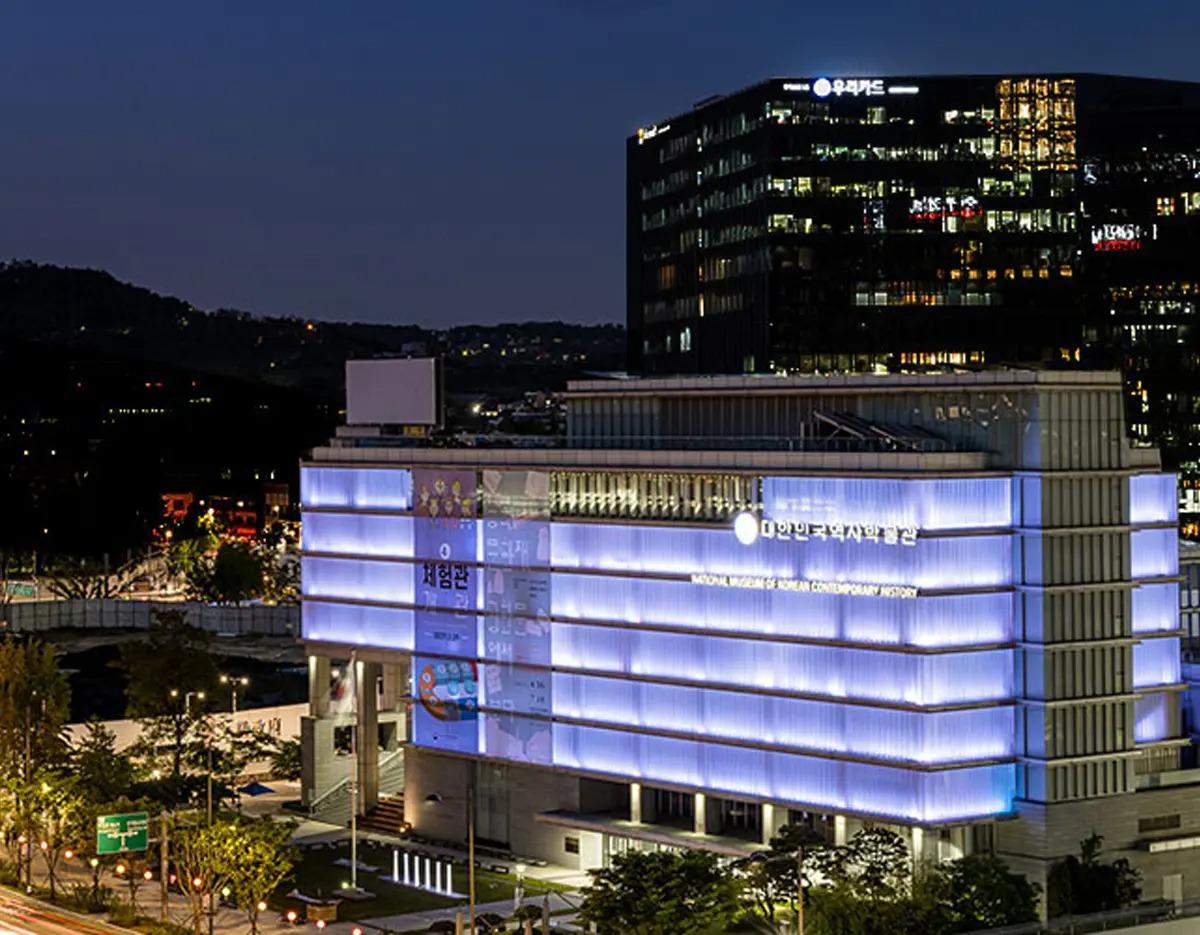 Night view of the illuminated National Museum of Korean Contemporary History building with blue horizontal light bands.