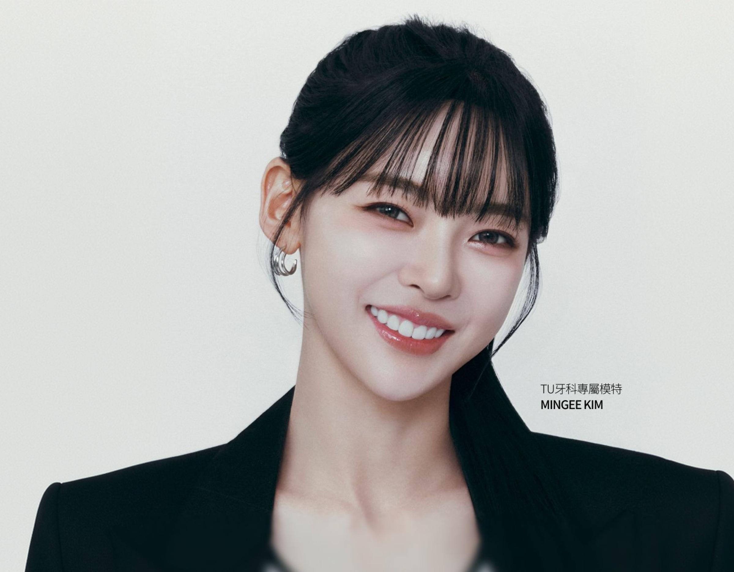 Young woman with dark hair and bangs, wearing a black blazer and hoop earring, smiling to show straight white teeth