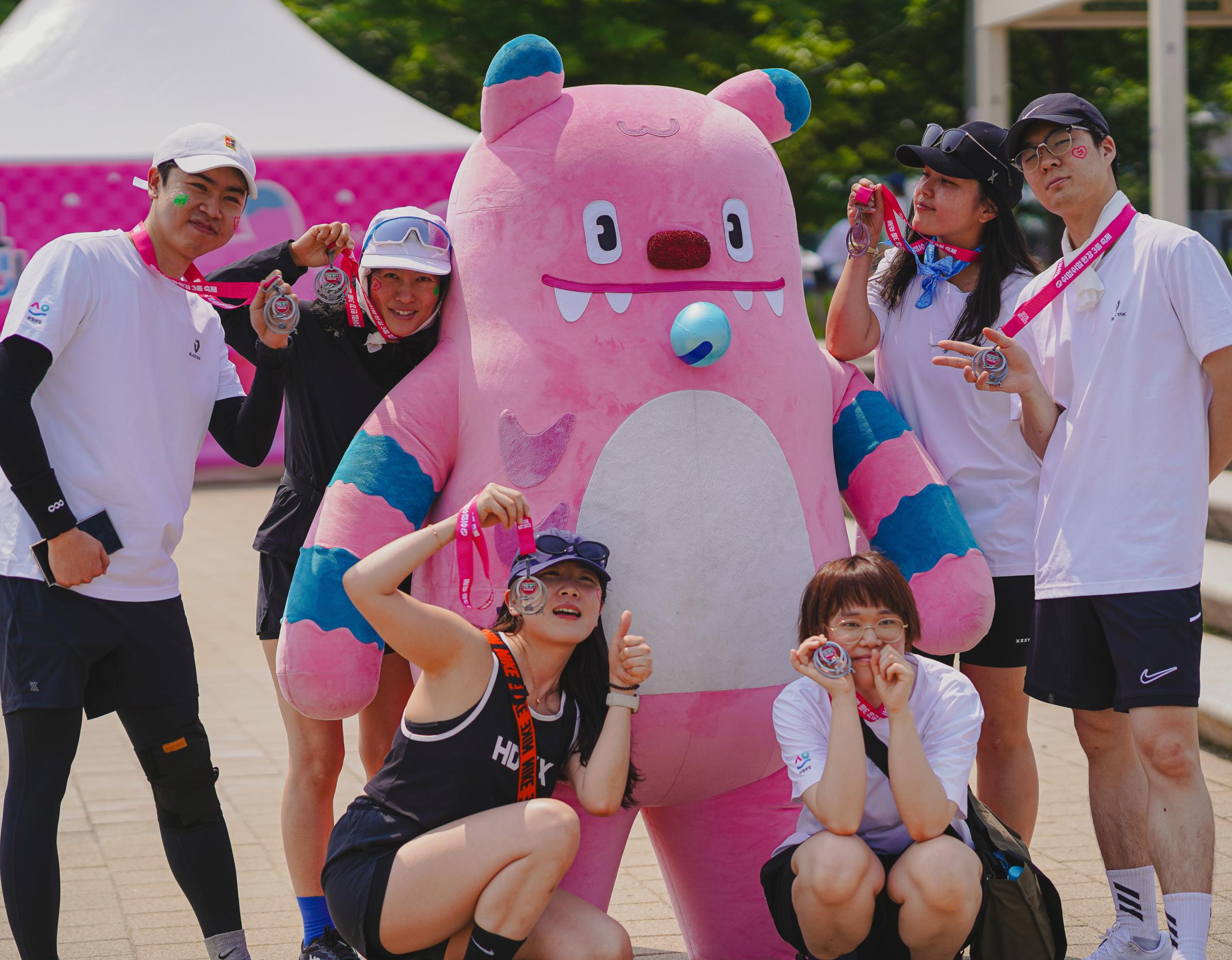 Pink mascot with blue-striped arms posing with masked participants holding medals at Han River triathlon festival