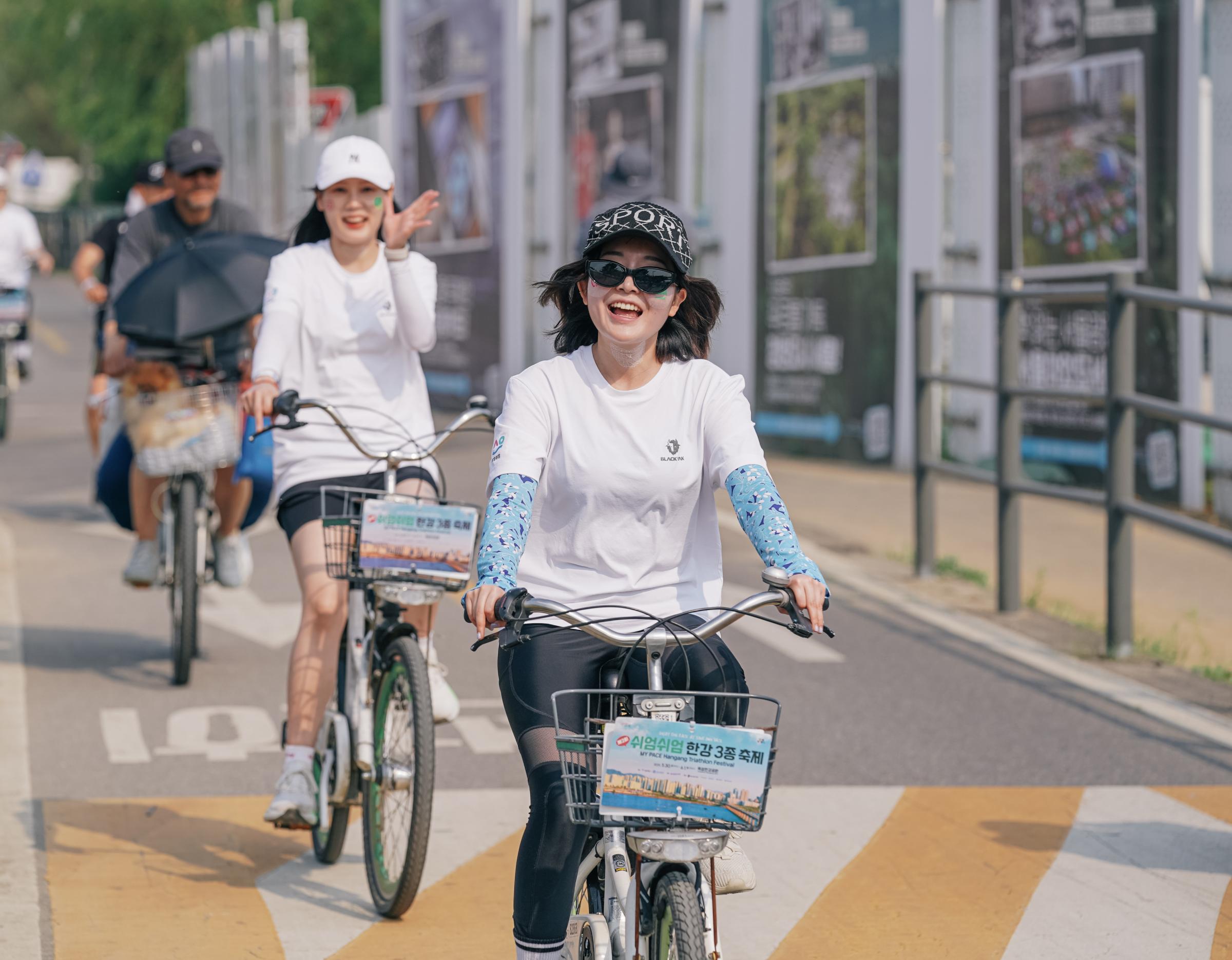 Woman in white T-shirt and patterned arm sleeves riding a rental bike with event sign, other cyclists behind on riverside path
