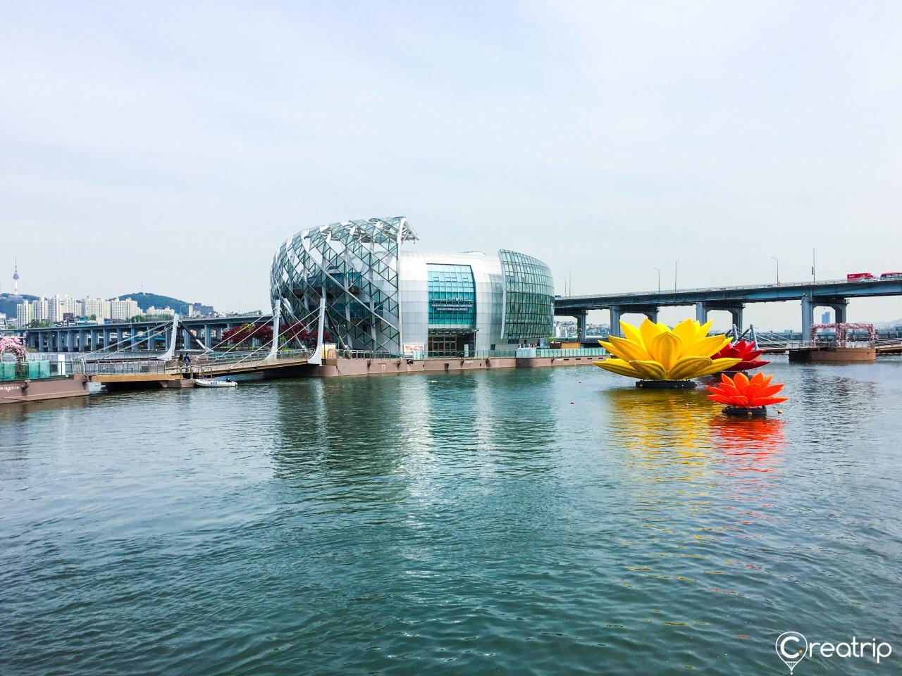 A panoramic view of the vibrant city of Seoul with landmarks like Sebit Island, towering buildings, and a serene lake surrounded by flowers. The sky is clear and blue with white clouds. Vehicles and people are enjoying leisure and recreation.