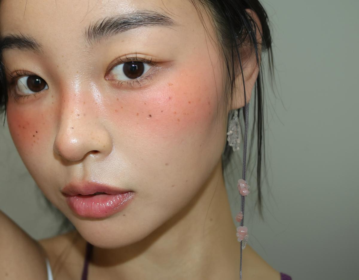 ORARE Cheongdam makeup close-up showing dewy skin, flushed rosy cheeks with faux freckles, glossy lips, and dangling beaded hair accessory.