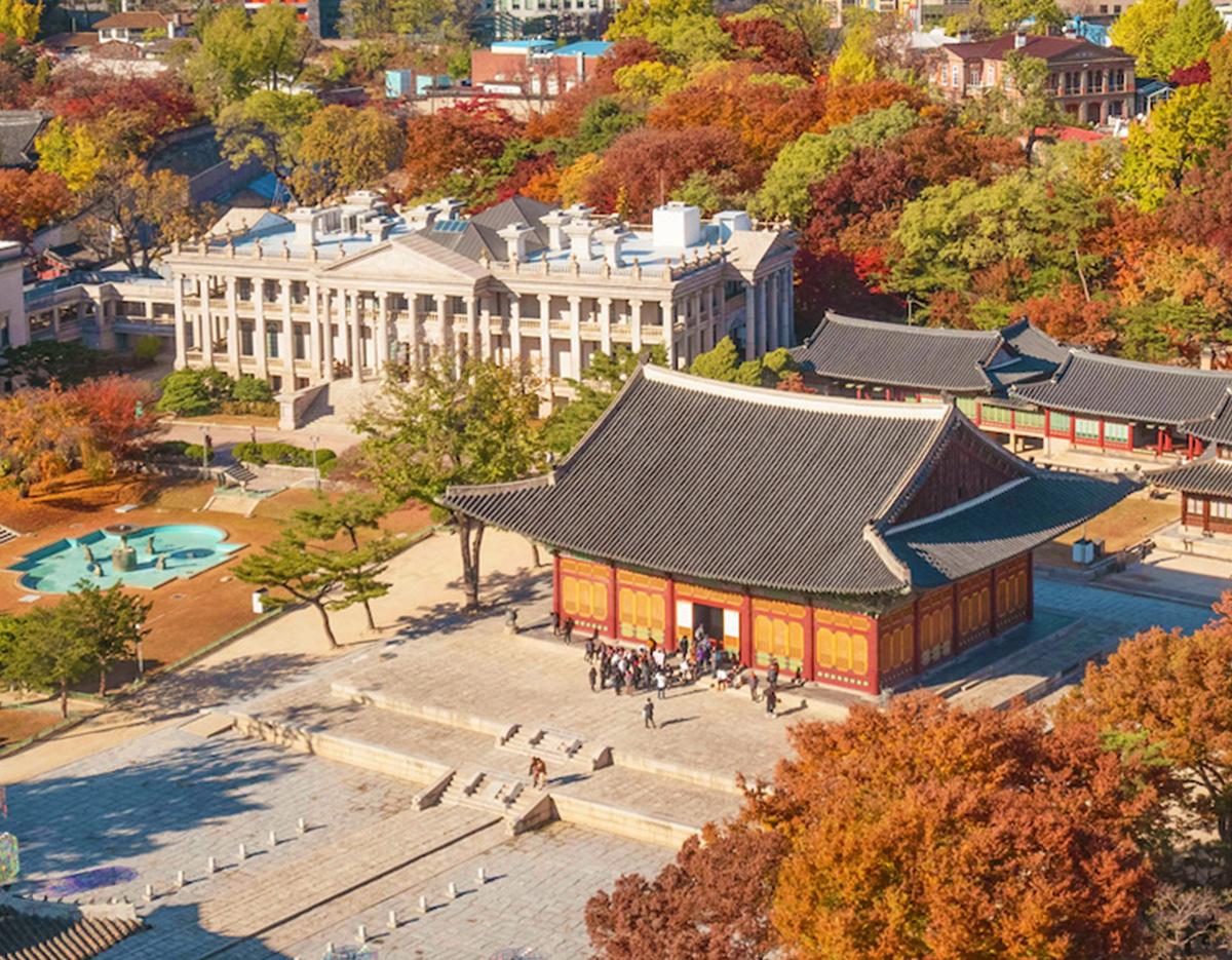 Gyeongbokgung pavilion with black tiled roof and crowds on stone courtyard, autumn trees and Western-style Seokjojeon building behind