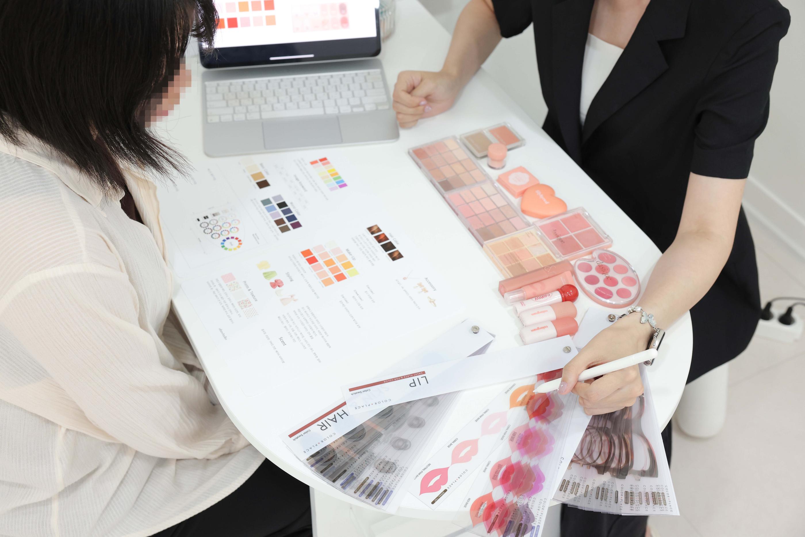 Consultant and client at white table reviewing personal color swatches, makeup palettes, lip and hair color guides, and laptop screen.