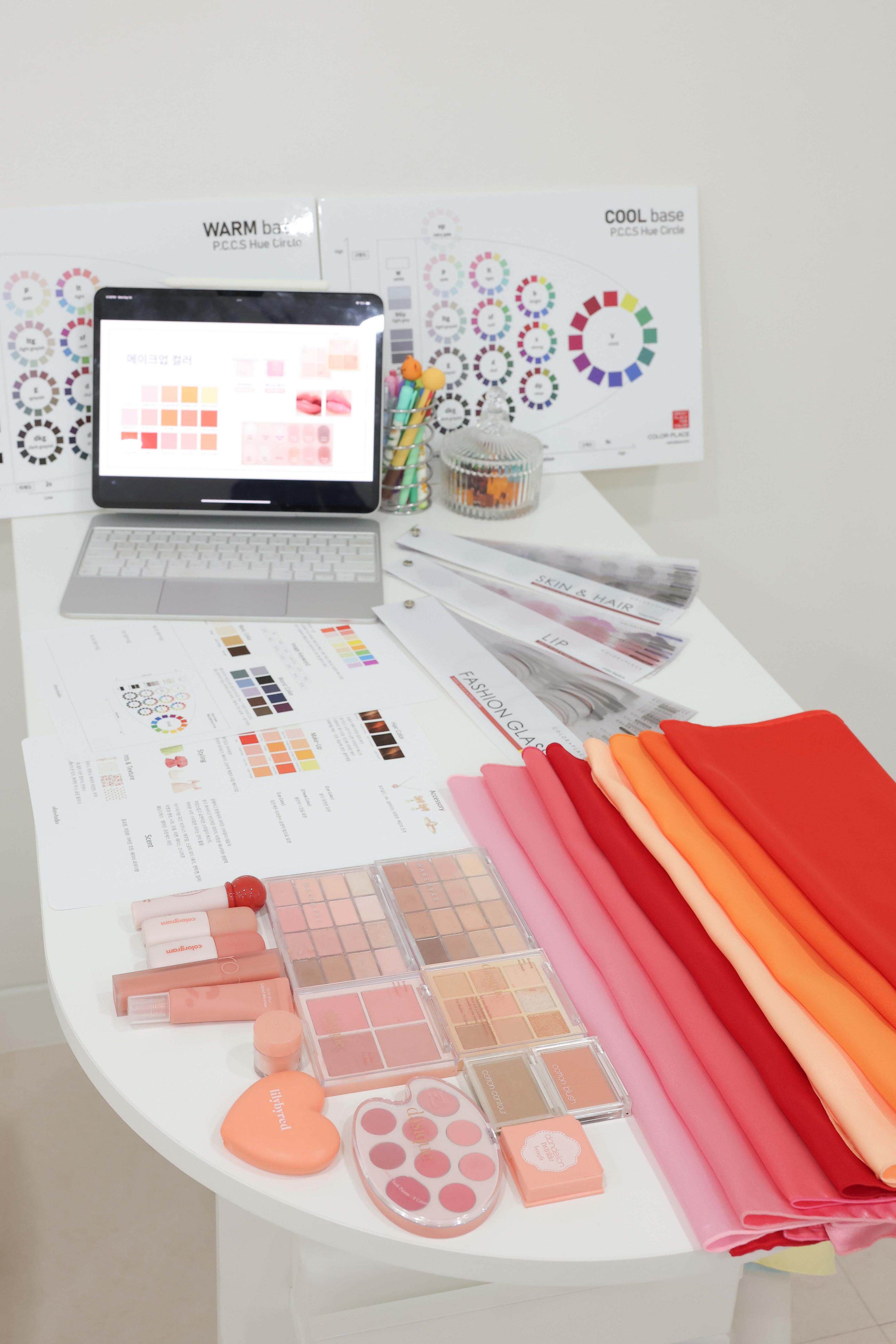 desk with color analysis tools: laptop showing swatches, warm/cool hue charts, makeup palettes, lip swatches, and folded pink-red drapes