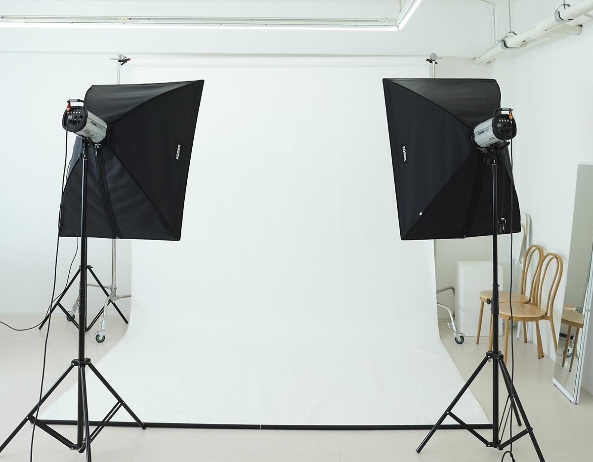 Minimal photography studio setup with white seamless backdrop, two softbox lights on stands and wooden chairs reflected in a standing mirror