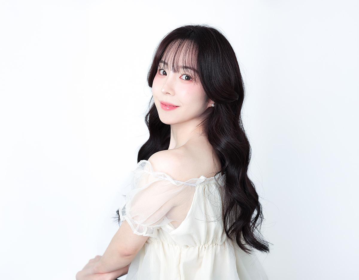 woman in off-shoulder sheer white dress turned three-quarters, long dark wavy hair against plain white background