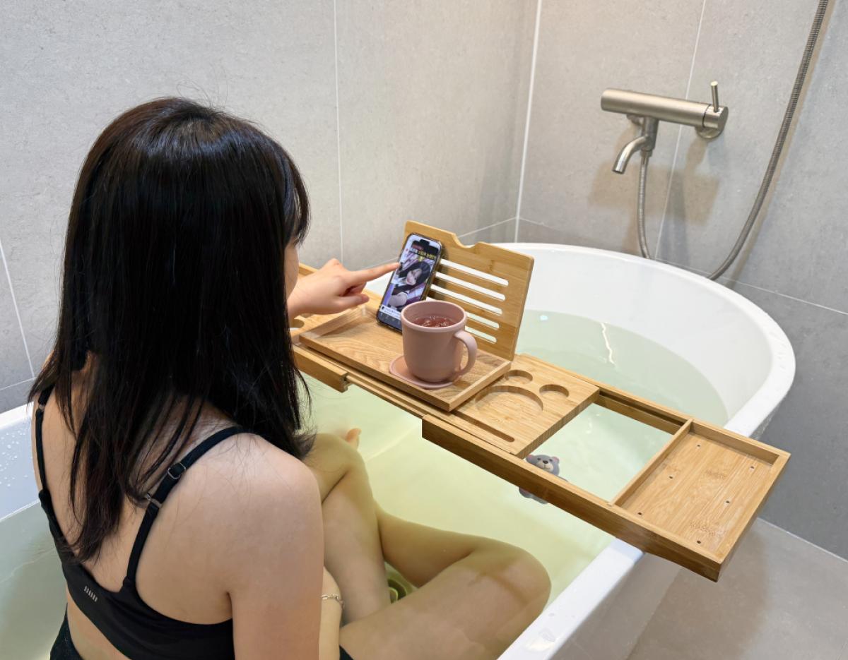woman sitting in a bathtub using a wooden bath caddy holding a smartphone and mug while pointing at the phone screen