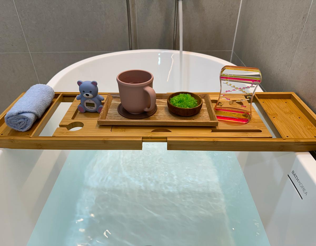 Wooden bath tray across a filled tub holding a rolled towel, toy bear timer, pink mug, bowl of green bath salts, and hourglass.