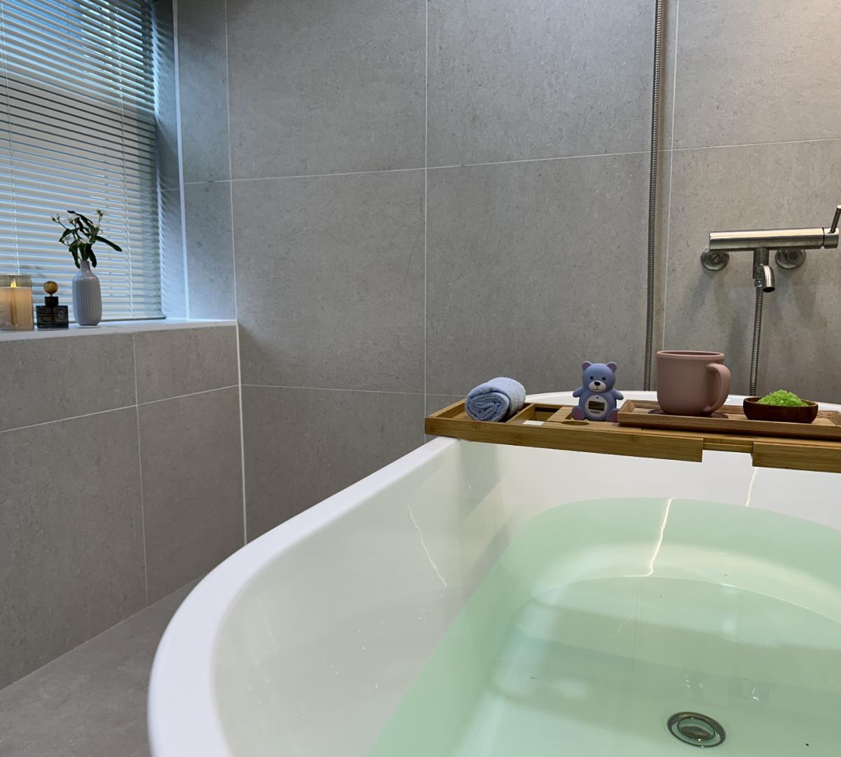 Minimal spa bathroom with a filled white bathtub, wooden bath tray holding a rolled towel, mug, small bear figurine and plant.
