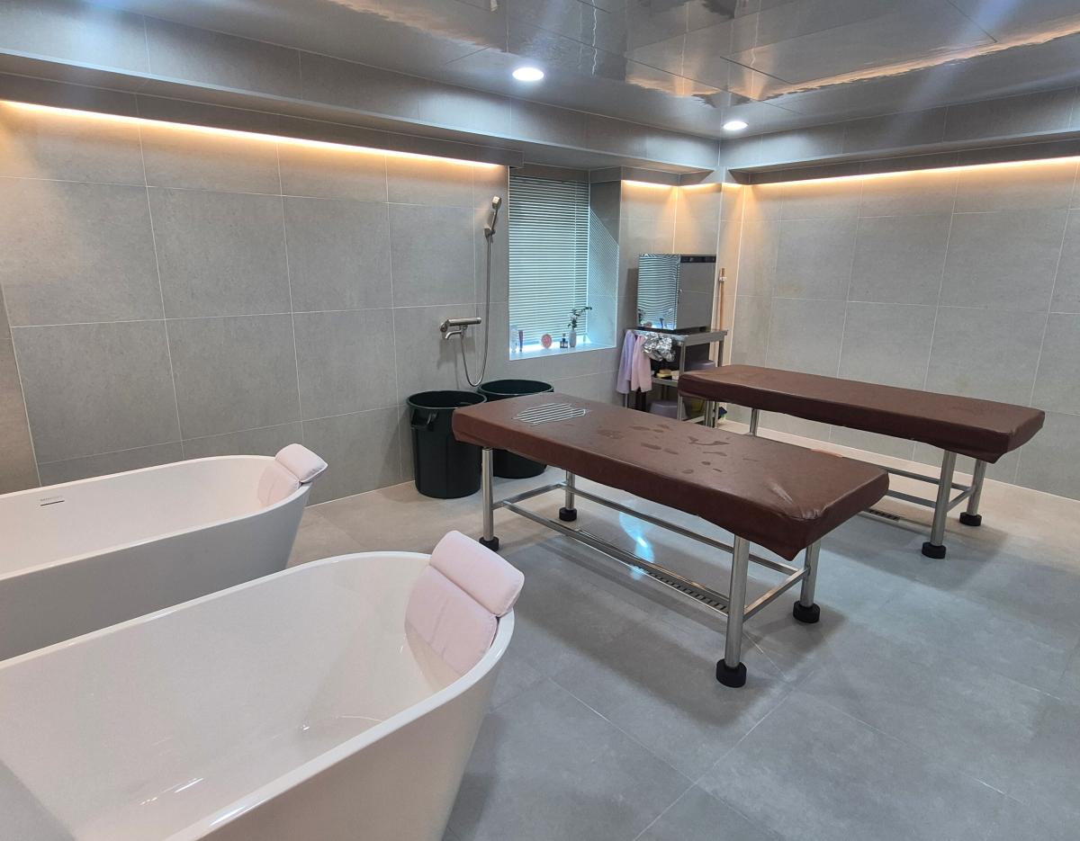 Women-only sesin room with two white soaking tubs with headrests and two brown massage tables in tiled space