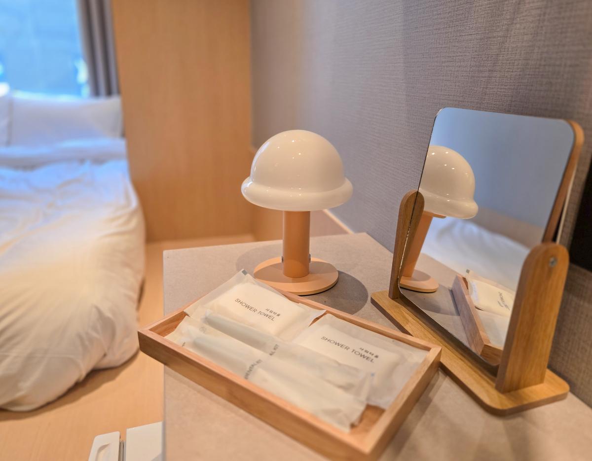 床頭櫃上木盤擺放數包標示「SHOWER TOWEL」的一次性毛巾，旁邊有圓頂小檯燈及木框座鏡，背景見白色床舖。