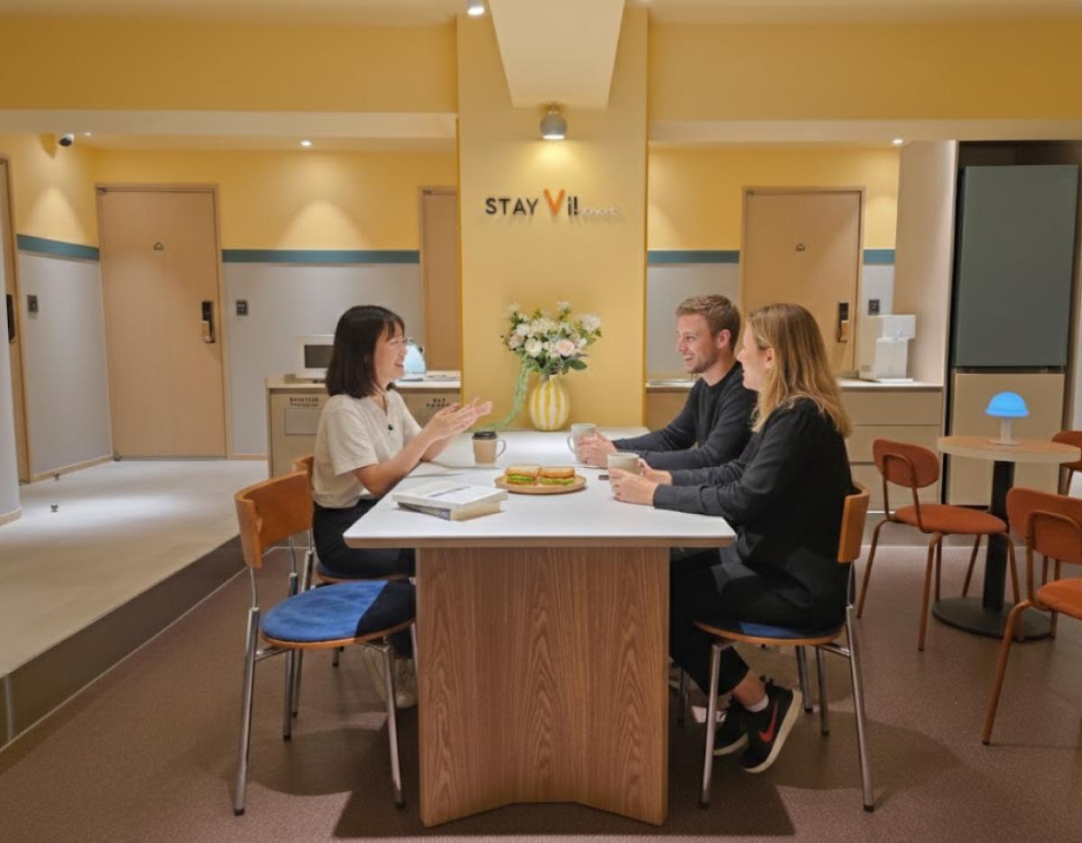 Stay Vi 鐘路公共休息區，黃色牆壁中央擺有花瓶，三位客人在長桌邊喝咖啡討論，周圍有座椅與房門背景