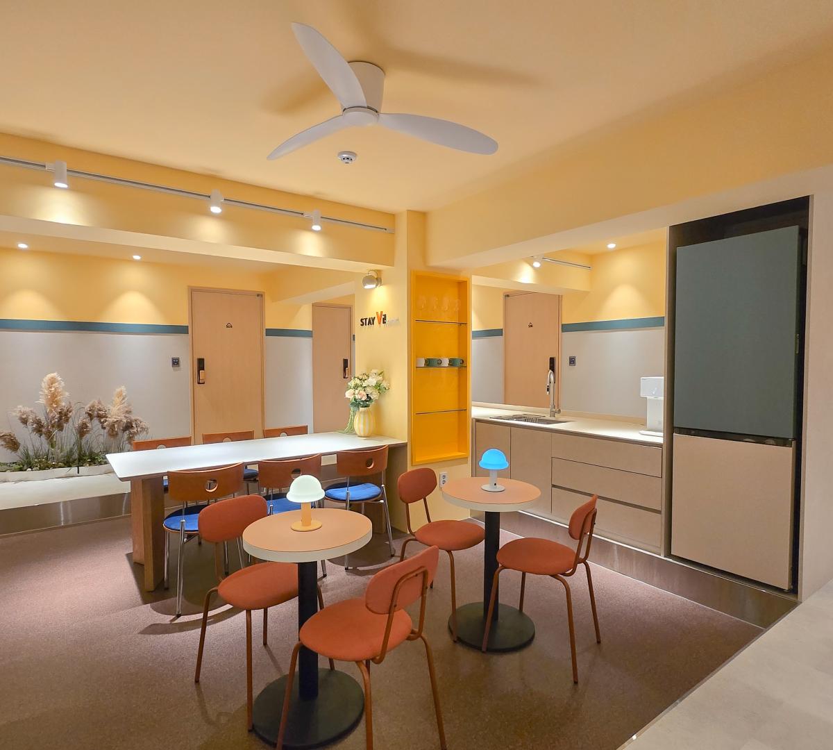 共用Lounge內景，暖黃色牆身和吊扇、長桌與多張橙色餐椅、小圓桌配彩色桌燈及開放式廚房區。