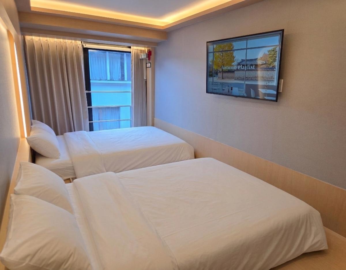 簡約雙床房內景，兩張鋪白床單的單人/雙人牀並排，牆上掛平面電視，窗邊有落地窗和窗簾，暖光天花燈。