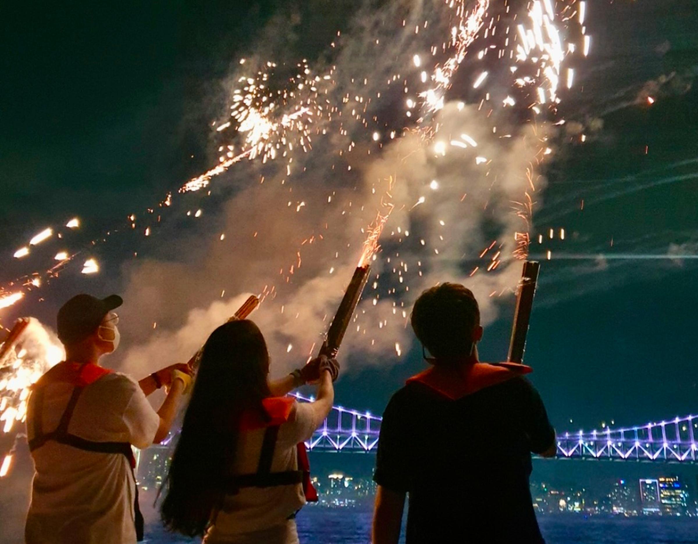 three people on a yacht at night firing handheld sparklers toward sky with illuminated bridge and city lights in background