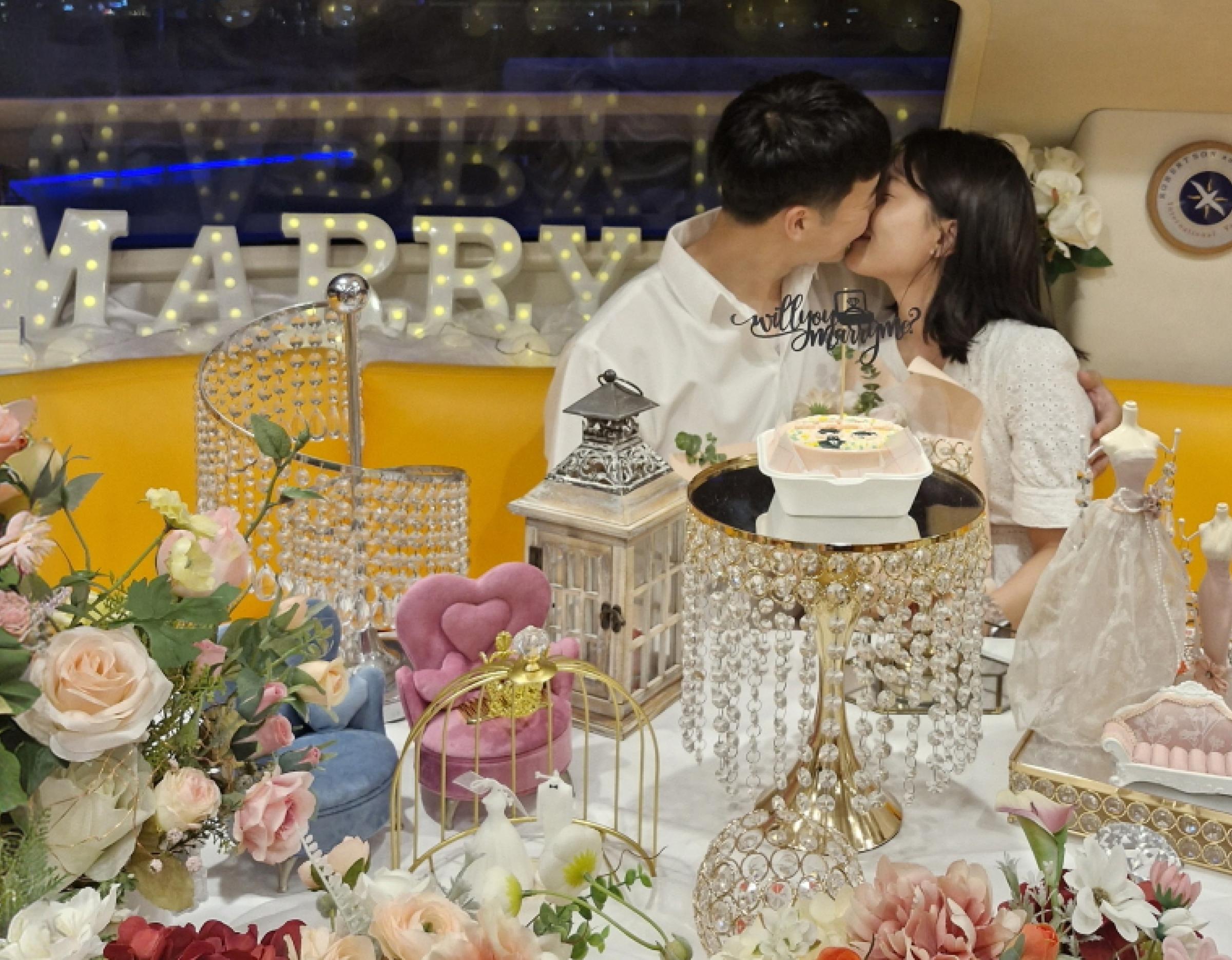 couple kissing behind a decorated proposal table with flowers, crystal cake stand, lantern and "MARRY" marquee lights