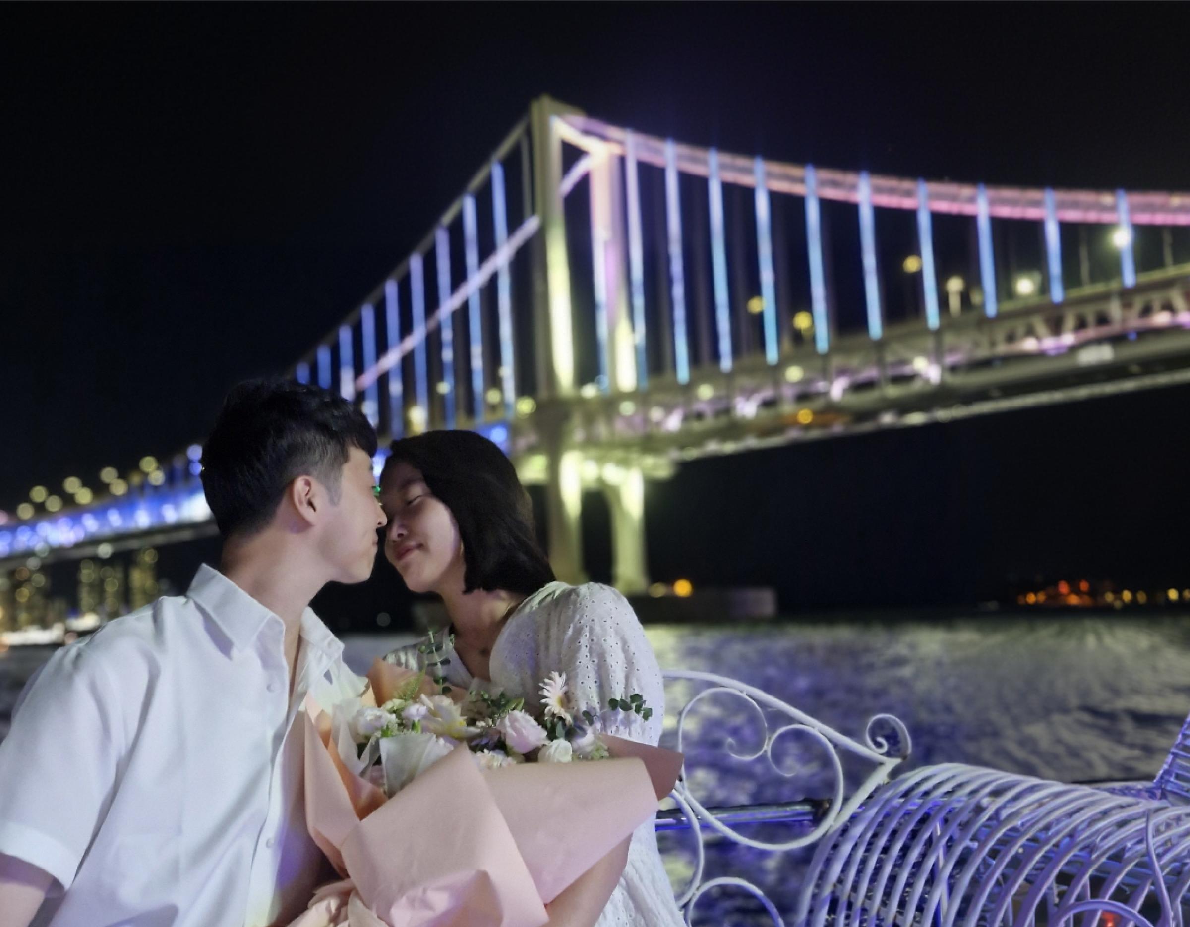 couple sharing a kiss on a yacht at night holding a bouquet with illuminated Gwangan Bridge in the background