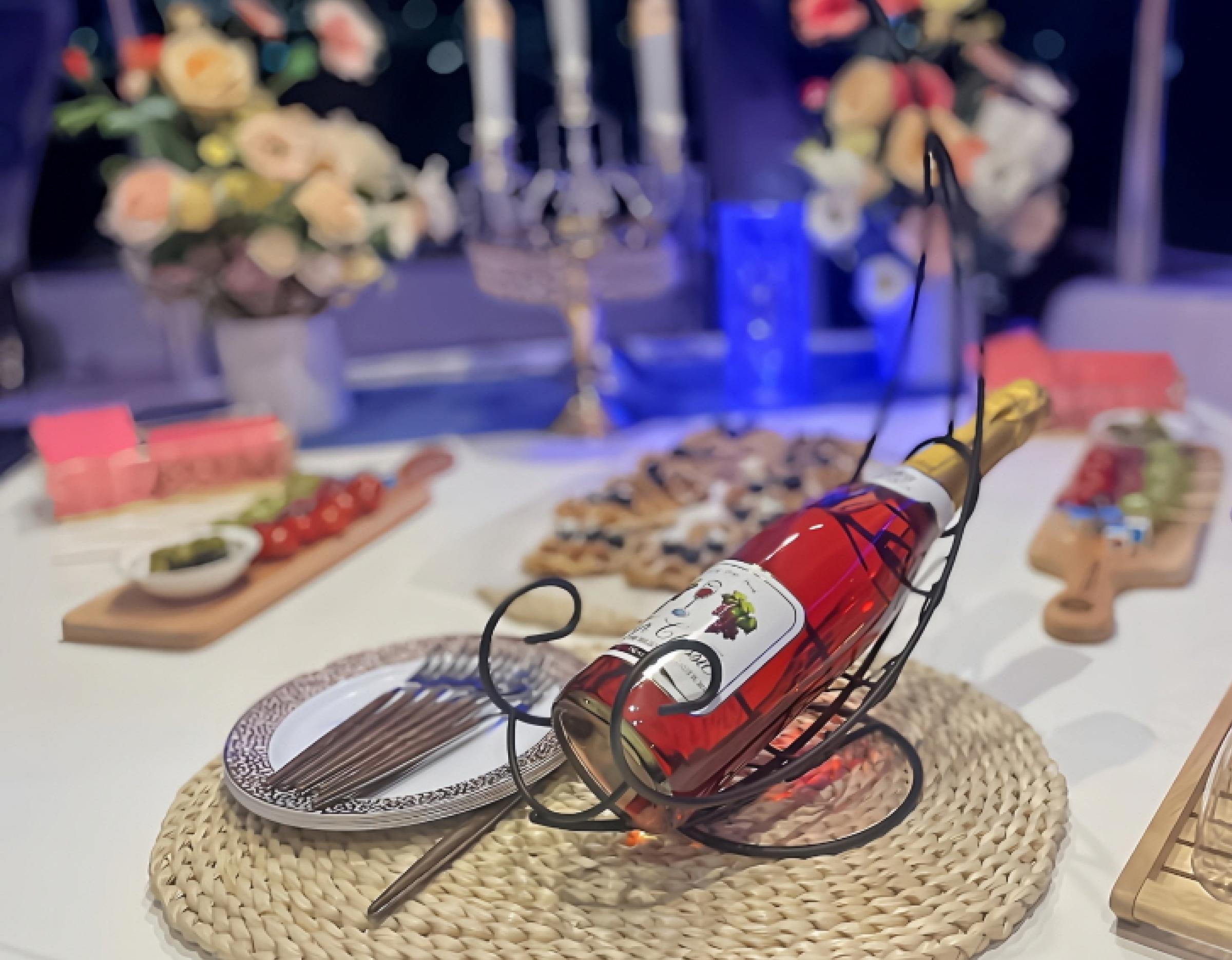 Bottle of sparkling rosé in a black metal cradle on woven placemat with plates, utensils and blurred floral table decor behind