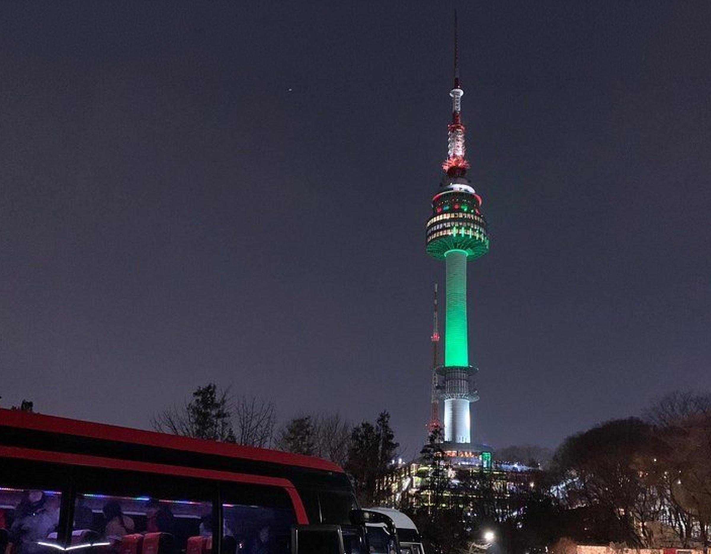 N Seoul Tower lit green and white at night rising above trees with a dark sky and a cable car bus in foreground