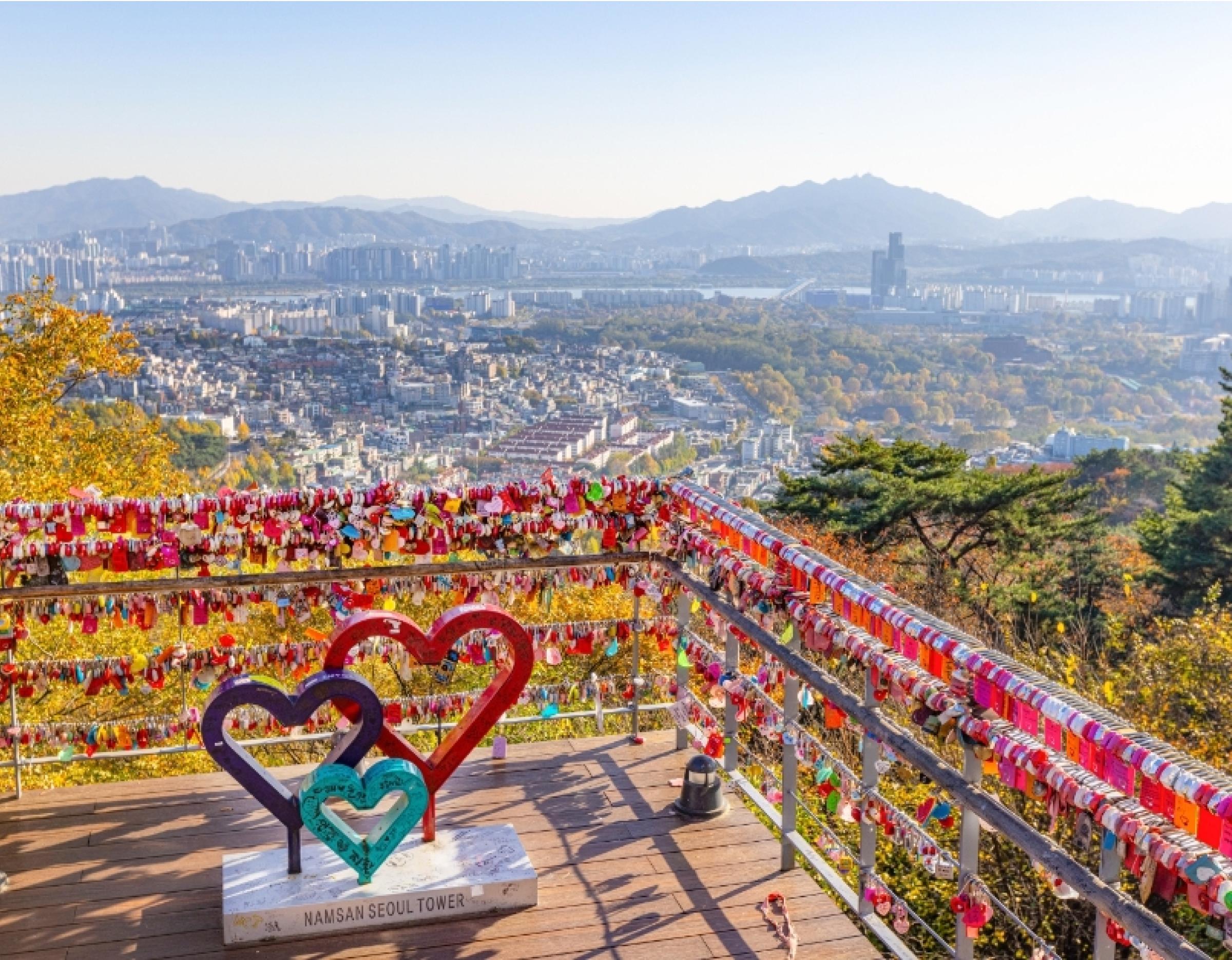 Colorful love locks covering railings at N Seoul Tower observation deck with heart sculptures and panoramic Seoul city view.