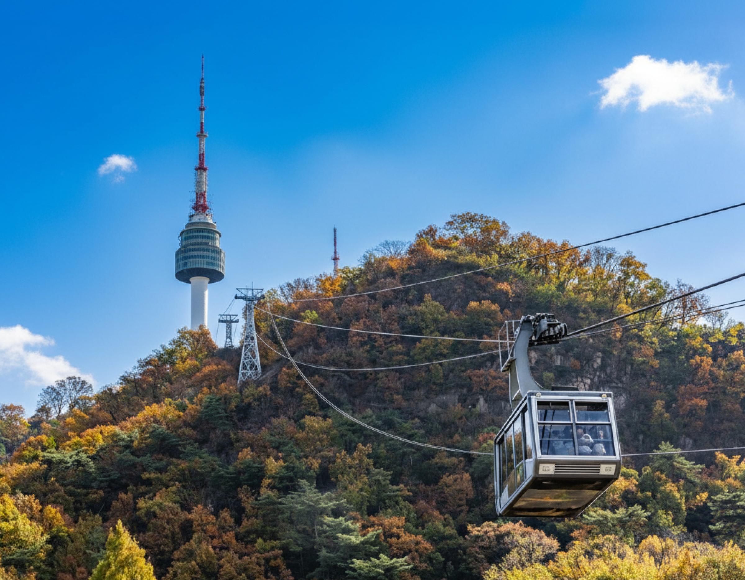 N Seoul Tower on wooded hill with autumn foliage and a cable car cabin suspended on cables approaching the tower