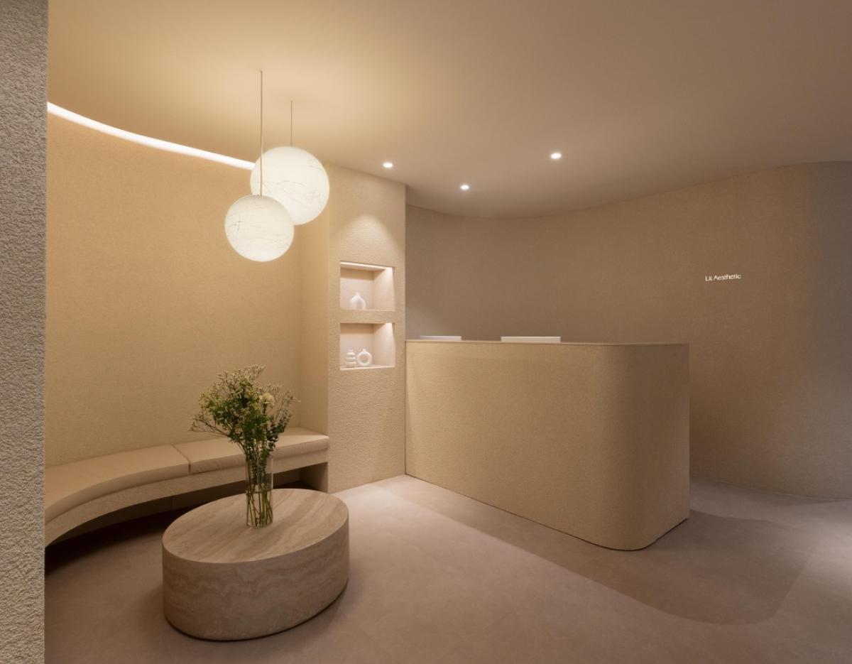 Minimal beige reception area with curved bench, oval stone coffee table with flowers, pendant globe lights and desk.