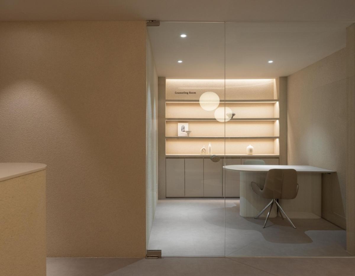 Minimalist consultation room with glass door, backlit shelves, round white table and beige swivel chair at Lit Aesthetic.