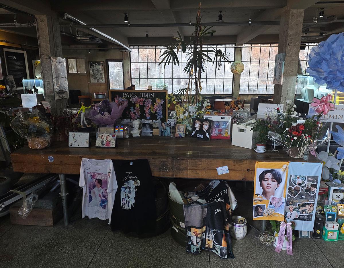 Indoor shrine table filled with Park Jimin fan gifts: flowers, photos, banners, shirts and memorabilia in a cafe-like space
